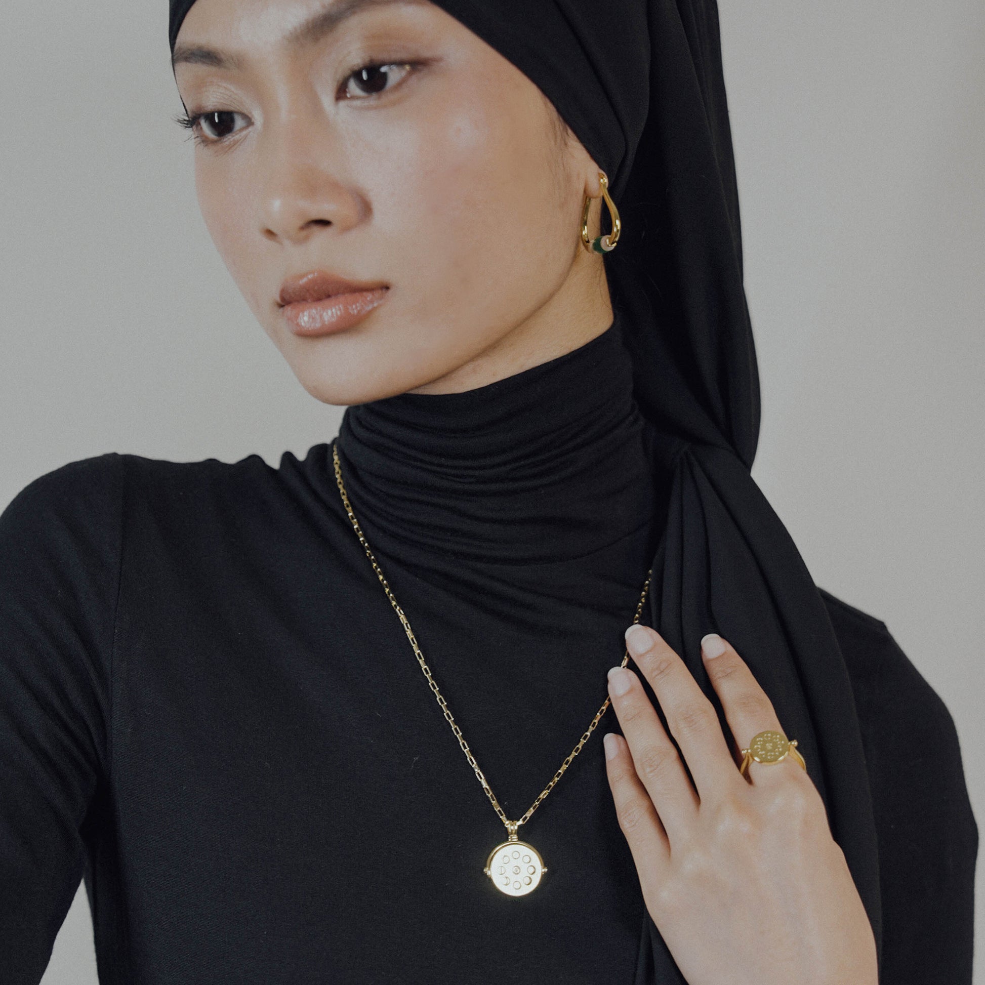 Woman wearing a black hijab and black turtleneck with gold jewelry on a plain background