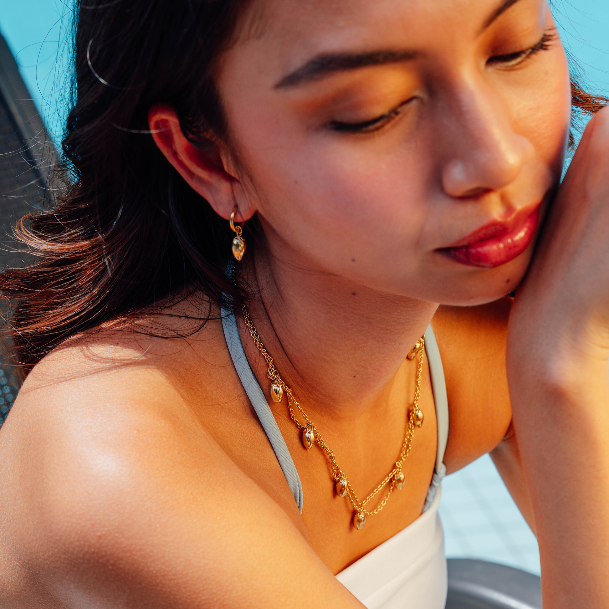 A close-up lifestyle shot of a woman wearing a gold necklace and matching drop earring, with warm sunlight highlighting her skin and the jewellery.