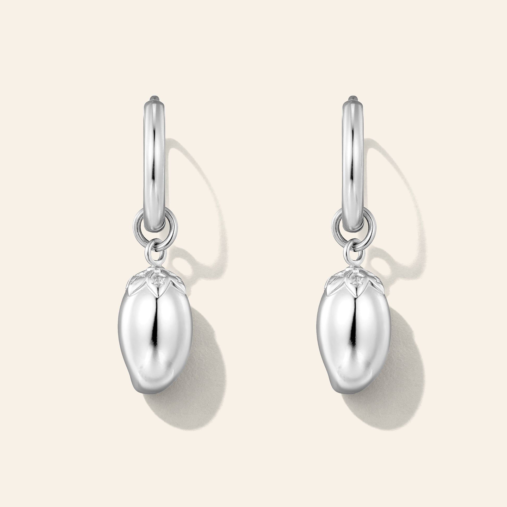 A pair of silver dangle earrings shaped like glossy, mango-shaped fruit charms, suspended from simple silver hoop posts, shown on a light beige background with soft drop shadows.