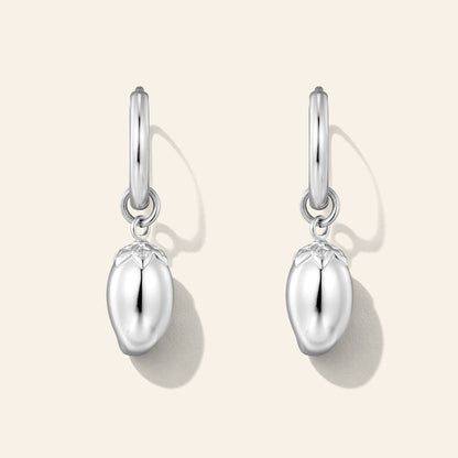 A pair of silver dangle earrings shaped like glossy, mango-shaped fruit charms, suspended from simple silver hoop posts, shown on a light beige background with soft drop shadows.