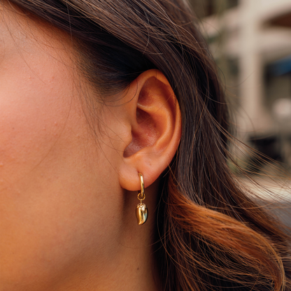 A close-up of a person's ear wearing a small gold hoop earring with a dangling mango charm.