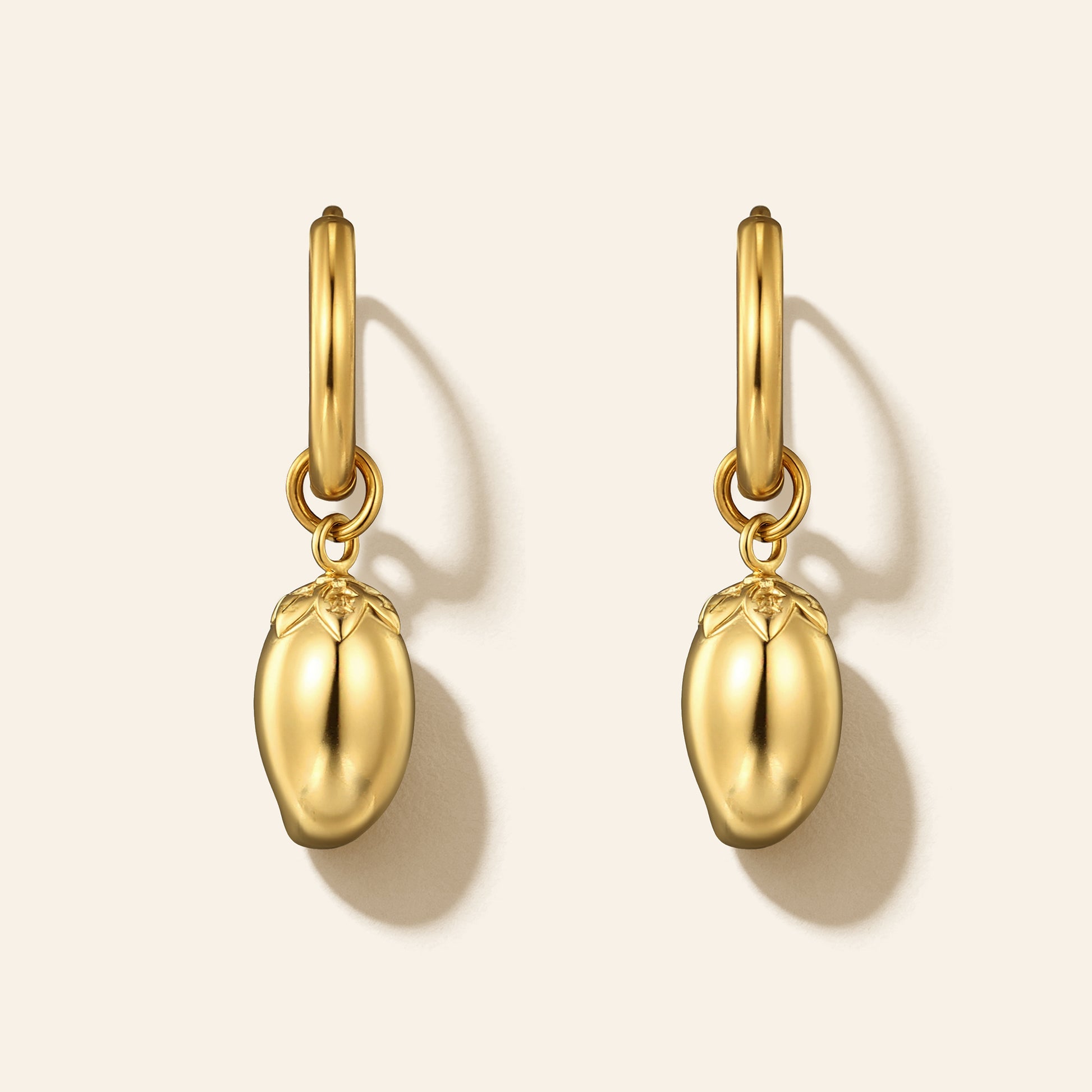 A pair of gold hoop earrings with dangling sculpted mango charms, shown on a light beige background with soft shadows.