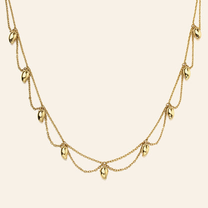 A gold multi-layered necklace featuring small, polished mango-shaped charms hanging from delicate chains against a solid cream background.