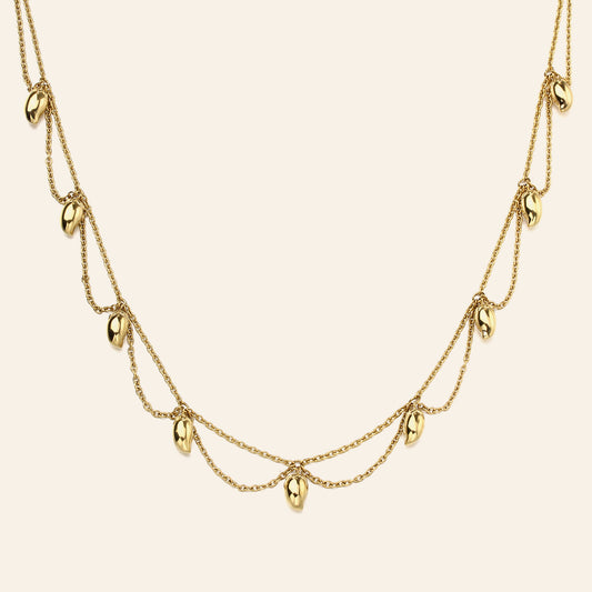 A gold multi-layered necklace featuring small, polished mango-shaped charms hanging from delicate chains against a solid cream background.