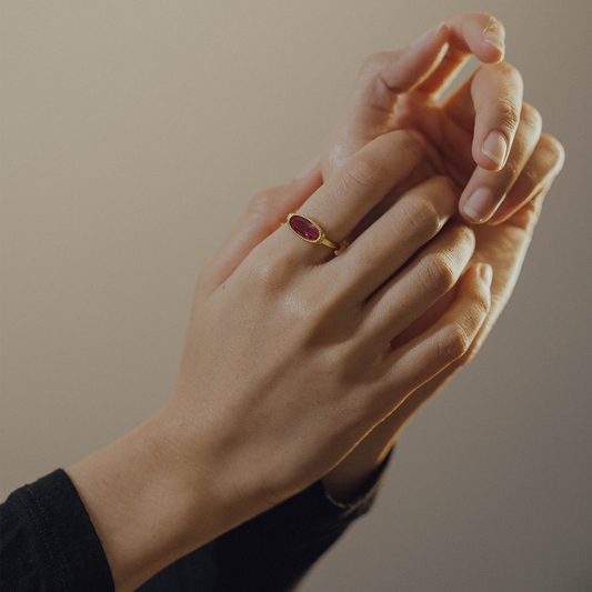 Close-up of a hand wearing a gold ring with a red gemstone on a neutral background