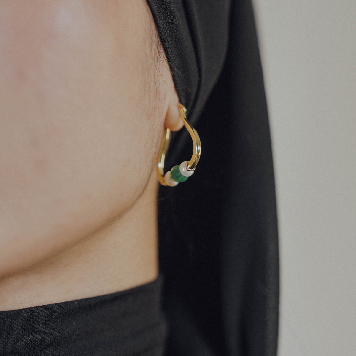 Gold hoop earring with green beads and pearl worn by a person against a neutral background