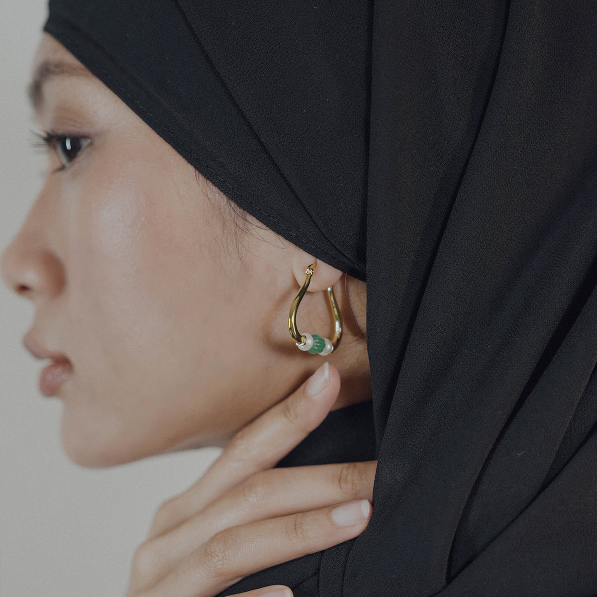 Woman wearing a gold earring with a green gemstone, partially covered by a black hijab.