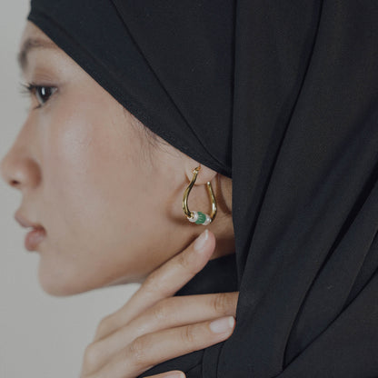 Woman wearing a gold earring with a green gemstone, partially covered by a black hijab.