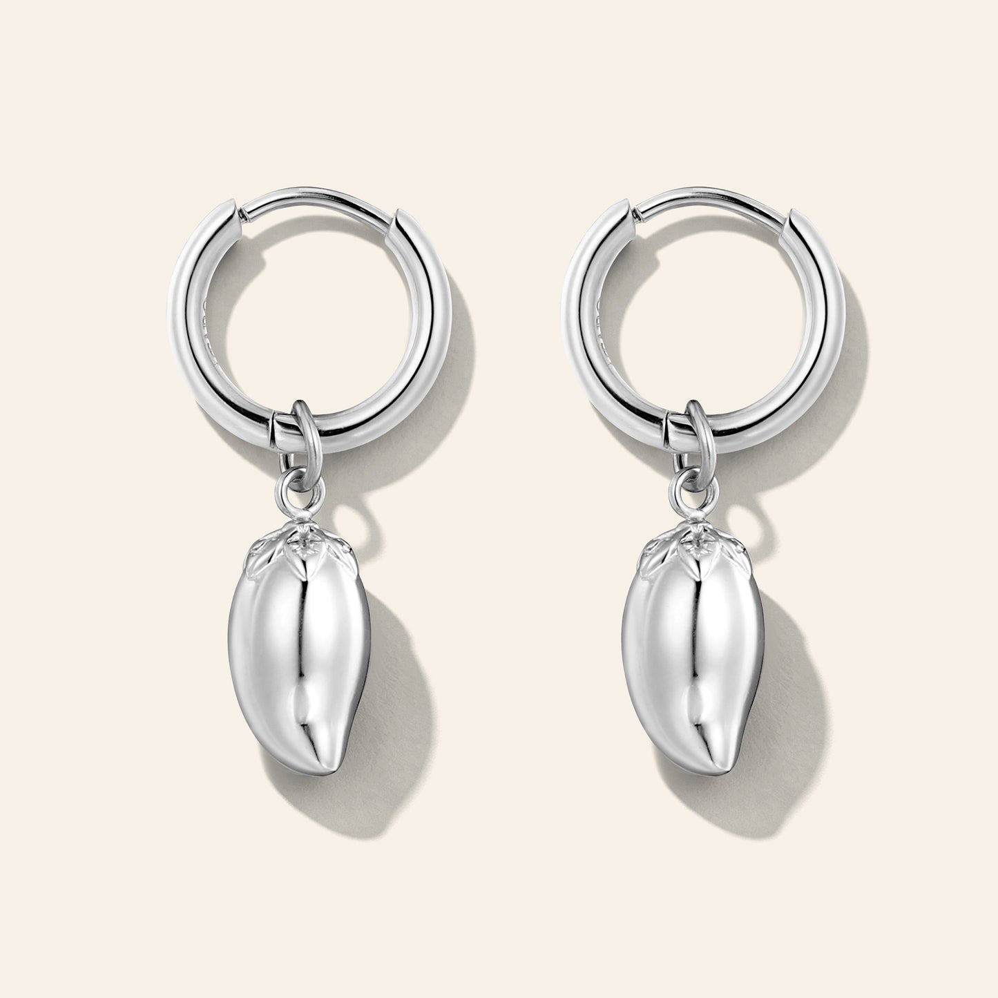 A pair of silver huggie hoop earrings with dangling polished metallic mango charms, displayed on a light beige background.