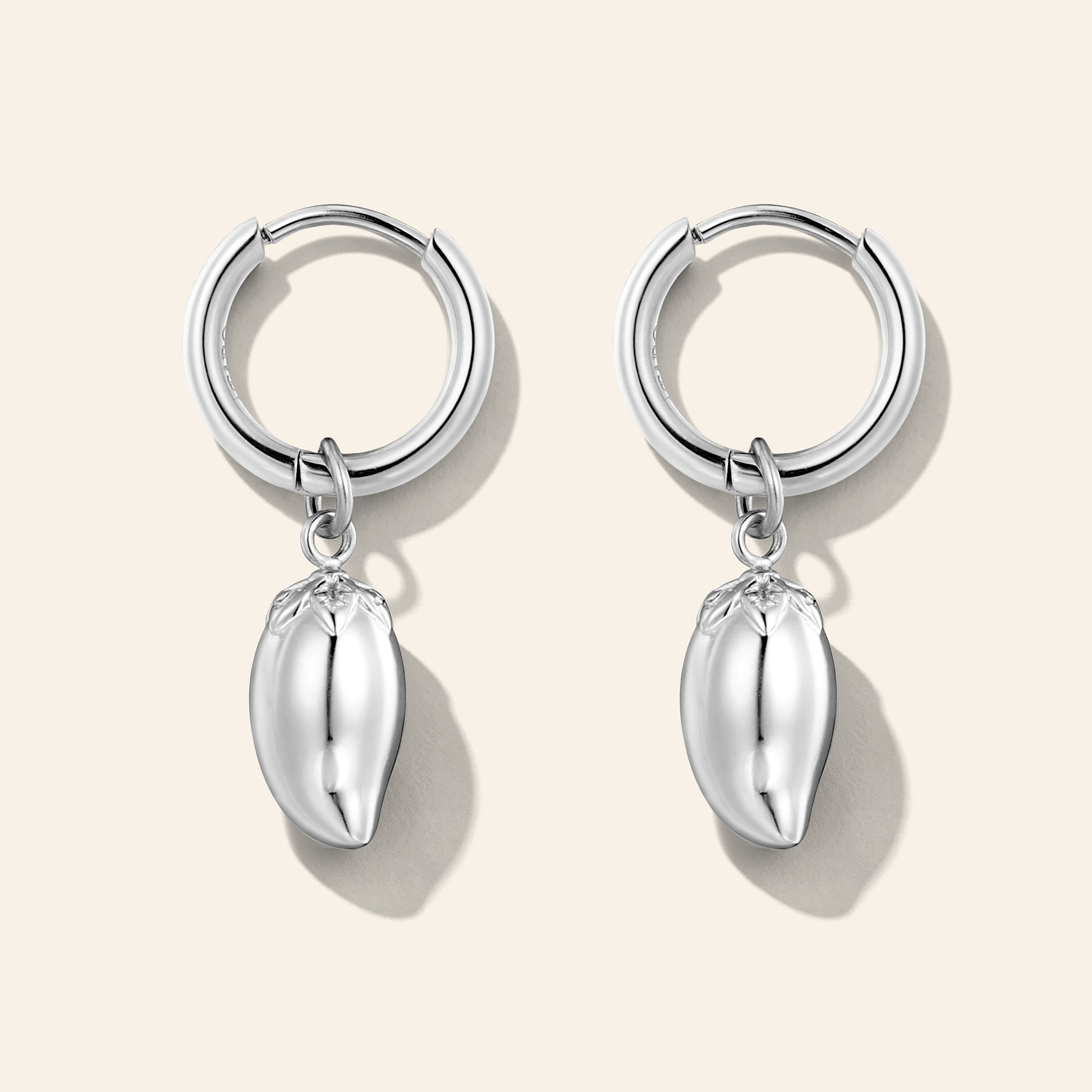 A pair of silver huggie hoop earrings with dangling polished metallic mango charms, displayed on a light beige background.