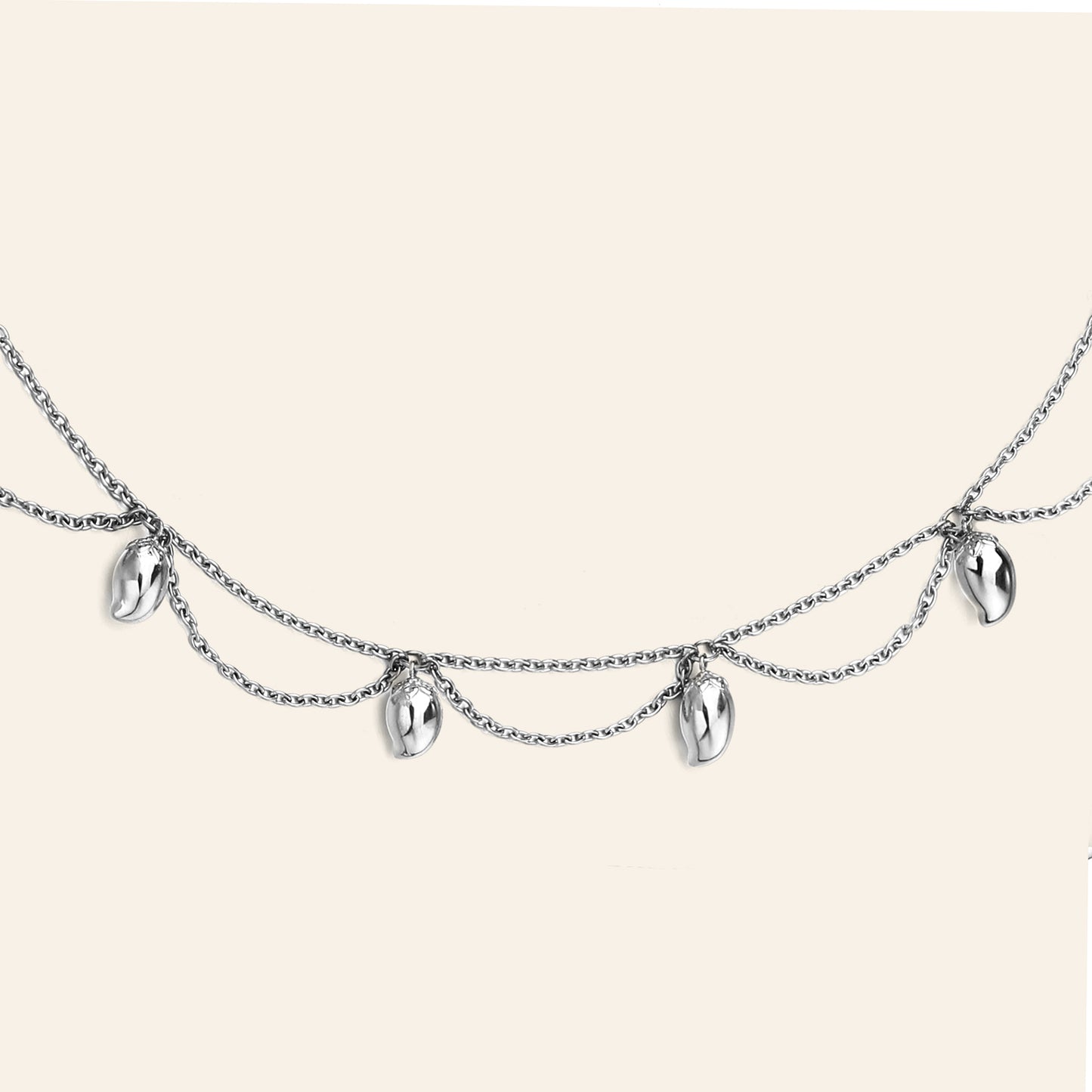 A silver double-layered chain necklace featuring five small, polished metallic mango shaped charms, displayed against a solid light beige background.