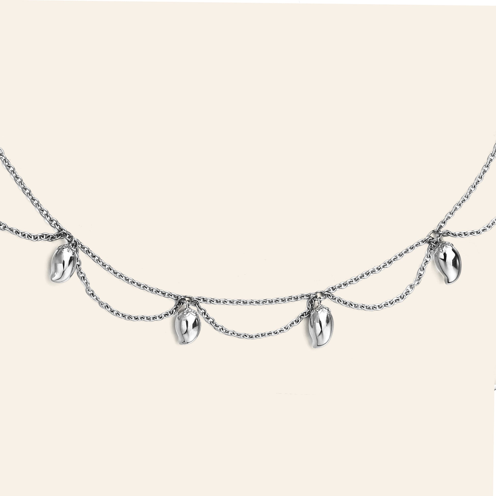 A silver double-layered chain necklace featuring five small, polished metallic mango shaped charms, displayed against a solid light beige background.
