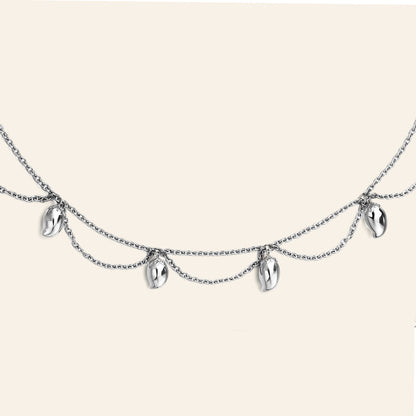 A silver double-layered chain necklace featuring five small, polished metallic mango shaped charms, displayed against a solid light beige background.