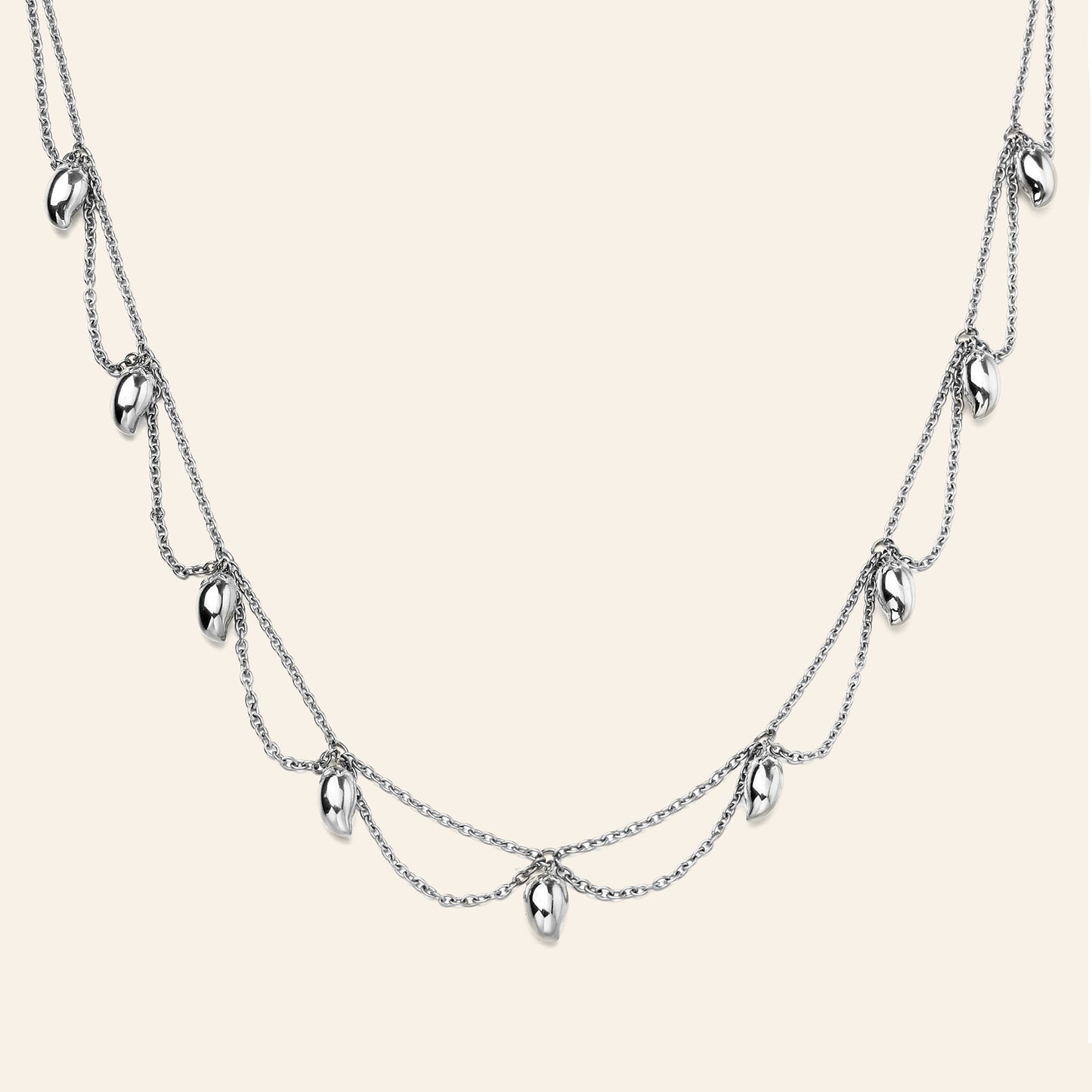 A silver multi-layered necklace featuring three draped chains with small mango shaped metallic charms, displayed against a solid light beige background.