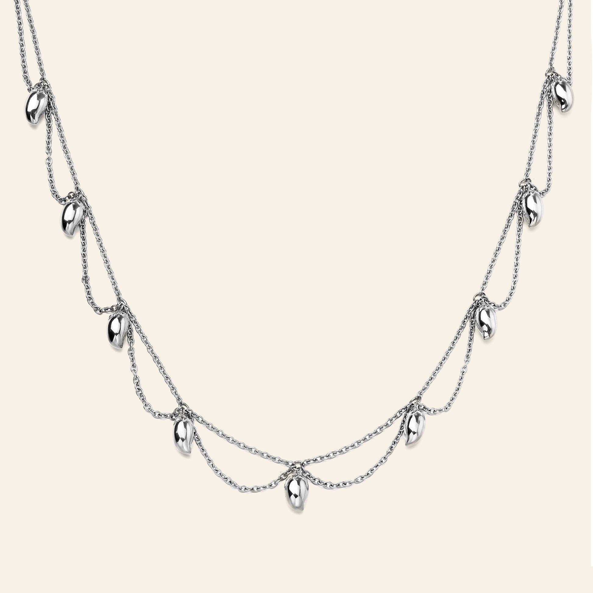 A silver multi-layered necklace featuring three draped chains with small mango shaped metallic charms, displayed against a solid light beige background.