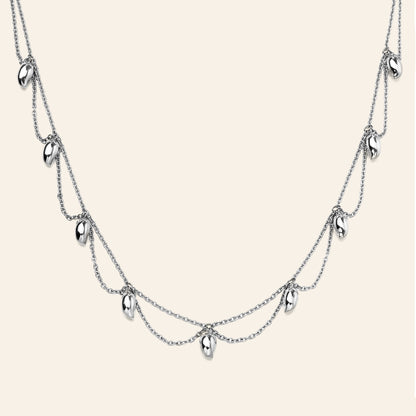 A silver multi-layered necklace featuring three draped chains with small mango shaped metallic charms, displayed against a solid light beige background.