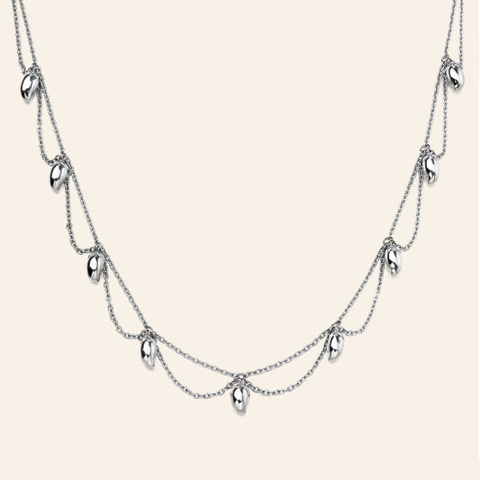 A silver multi-layered necklace featuring three draped chains with small mango shaped metallic charms, displayed against a solid light beige background.