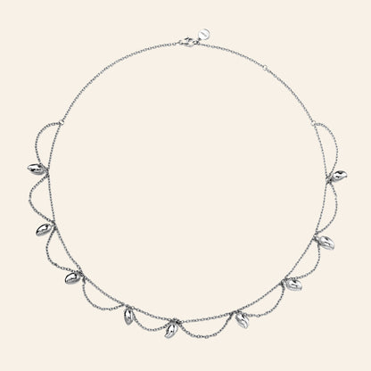 A delicate silver-toned necklace with a fine chain, draped mango chains, and small mango-shaped clear stones, displayed on a solid light beige background.