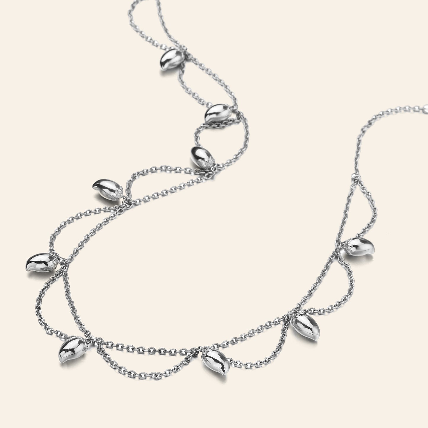 A silver double-strand necklace featuring dangling mango-shaped seed pods, displayed on a light cream background.