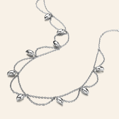 A silver double-strand necklace featuring dangling mango-shaped seed pods, displayed on a light cream background.